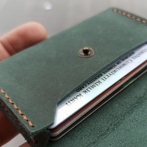 以下が含まれることがあります： オレンジ色のステッチと金属製のボタン留めが付いた緑色の革製のカードホルダー。ホルダーが開いており、トルコの身分証明書を含むいくつかのカードが入っています。