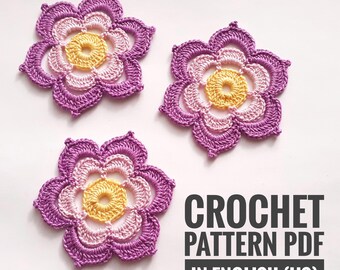 May include: Three handmade crochet flowers in shades of purple, pink, and yellow. The flowers have a layered petal design and a textured appearance. The text "CROCHET PATTERN PDF IN ENGLISH (US)" is displayed in the lower right corner.