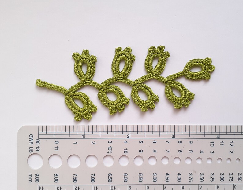 May include: A handmade, olive green crochet leaf with a delicate floral design. The leaf measures approximately 4 inches in length. The intricate details of the crochet work are visible, showcasing the craftsmanship. This item is perfect for embellishing clothing or craft projects.