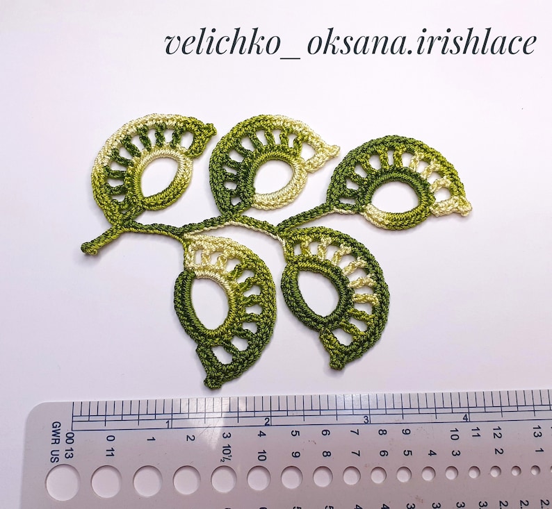 May include: A green and white crocheted leaf motif with a delicate, lacy design. The motif is approximately 4 inches wide and 3 inches tall.
