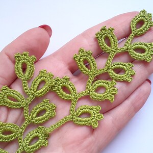 May include: A handmade, green crocheted leaf trim. The trim features a delicate design with oval-shaped leaves and small, bud-like details. The trim is shown against a light background, highlighting the intricate craftsmanship.