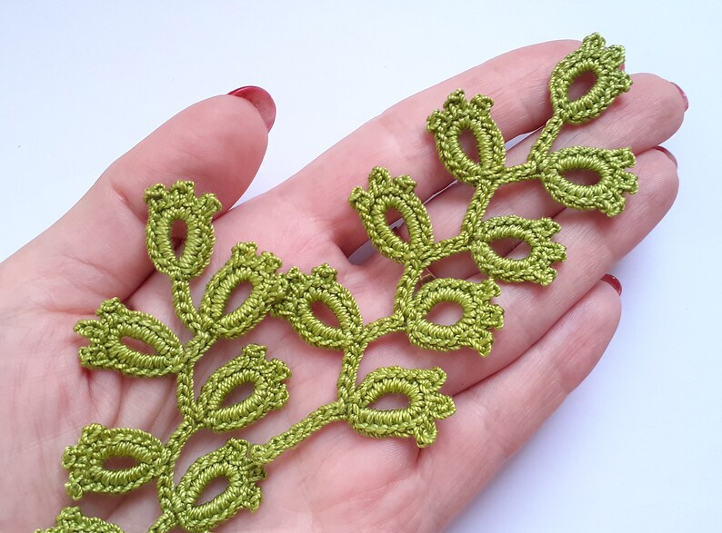 May include: A handmade, green crocheted leaf trim. The trim features a delicate design with oval-shaped leaves and small, bud-like details. The trim is shown against a light background, highlighting the intricate craftsmanship.
