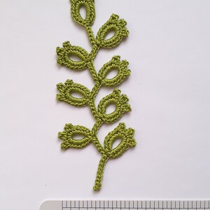 May include: A handmade, olive green crochet leaf with a stem and several flower-like buds. The leaf is placed on a white surface next to a ruler with inch measurements. The crochet work is detailed and intricate.