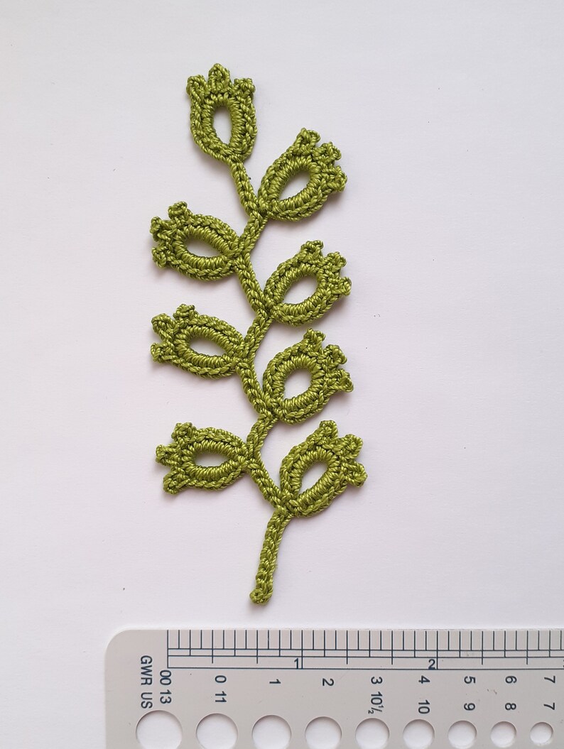 May include: A handmade, olive green crochet leaf with a stem and several flower-like buds. The leaf is placed on a white surface next to a ruler with inch measurements. The crochet work is detailed and intricate.