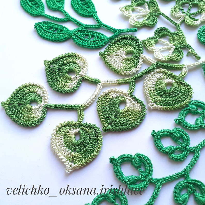 May include: A close-up of a crocheted green and white leaf design. The leaves are connected by a thin crocheted stem. The design is intricate and delicate.