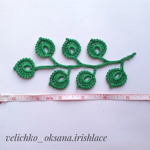 May include: Green crocheted leaf motif with a stem and six leaves. The motif is approximately 5 inches long and is measured with a white tape measure.
