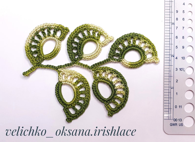May include: A crocheted green and cream leaf design with a ruler for scale. The design is made of multiple leaves connected by a stem. The leaves are in a gradient of green and cream.
