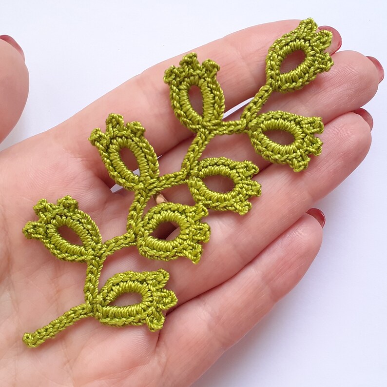 May include: A handmade, olive green crochet leaf with a stem and several oval-shaped leaves. The delicate, openwork design is visible, showcasing the intricate stitching. The leaf is likely a decorative element for crafts or embellishments.