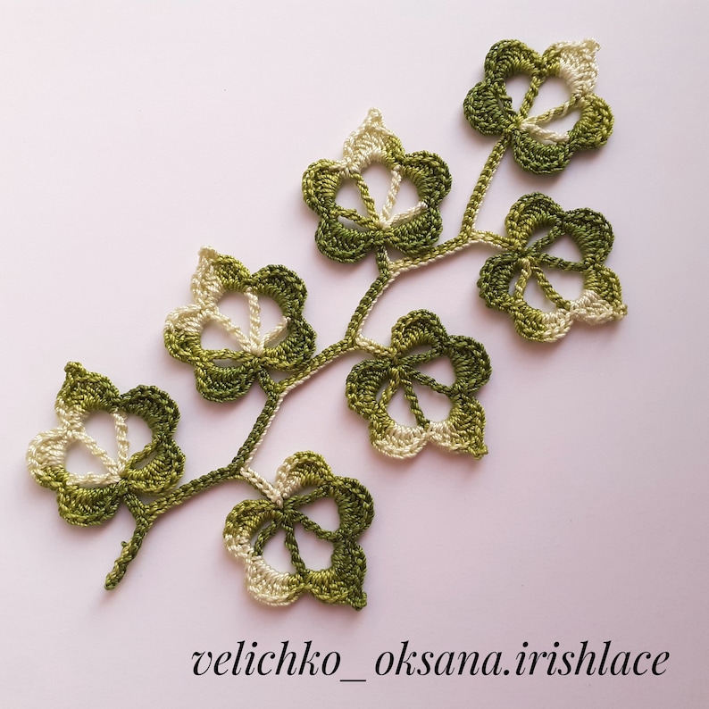May include: A crocheted green and white leaf design with a delicate, intricate pattern. The leaves are connected by a thin stem, creating a branch-like shape.