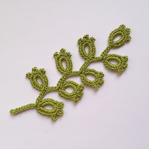 May include: A handmade, olive green crochet leaf with a delicate, openwork design. The leaf features multiple oval-shaped openings and small, bud-like details at the ends of the stems. The craftwork is set against a plain white background.