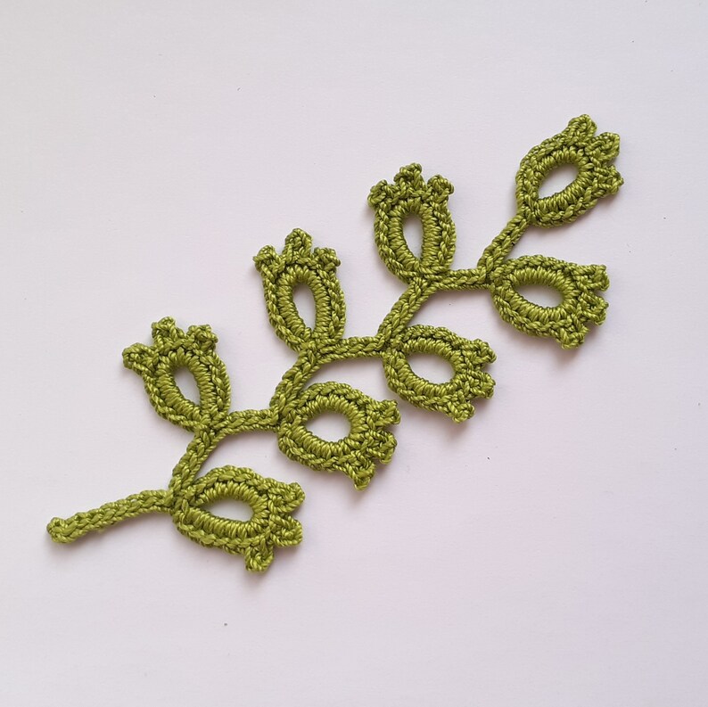 May include: A handmade, olive green crochet leaf with a delicate, openwork design. The leaf features multiple oval-shaped openings and small, bud-like details at the ends of the stems. The craftwork is set against a plain white background.