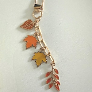 May include: A gold-toned metal keychain featuring a lobster clasp attached to a chain link. The keychain is adorned with four enamel charms: a small textured leaf, a solid orange maple leaf, a solid yellow maple leaf, and a branch with small orange leaves.