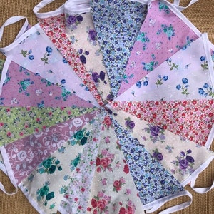 May include: A bunting banner with 12 triangular flags. Each flag is made of a different floral fabric in shades of pink, blue, and green. The flags are sewn together with white fabric.