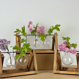 May include: Wooden display stands holding glass bulb vases with flowers. The vases are arranged in sets of one, two, and three, each with a different type of flower. The flowers include pink and purple blooms with green leaves.