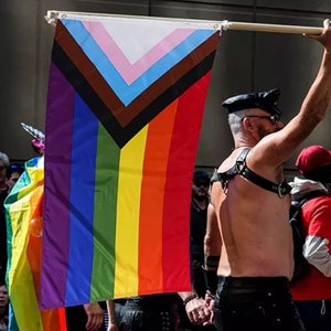 Può includere: Una persona tiene in mano una bandiera arcobaleno del pride con un design a chevron nero e marrone. La bandiera è fatta di tessuto e ha una striscia bianca, rosa, blu chiaro, blu scuro, verde, gialla, arancione e rossa.