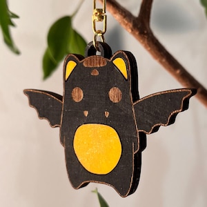 May include: Wooden bat-shaped keychain with a gold-colored clasp. The bat has a black body with yellow accents on the ears, belly, and wing tips. The keychain is hanging from a tree branch with green leaves.