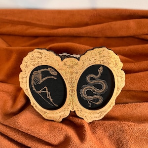 May include: A decorative wooden piece with two oval frames, each containing a black background. One frame features a skeleton, the other a snake. The frames are gold-colored with ornate detailing.