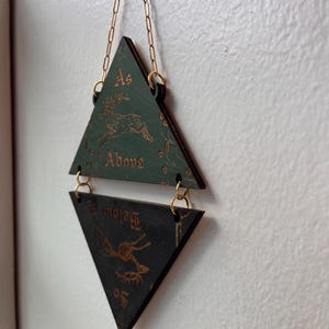 May include: A hanging wall decoration consisting of two triangular wooden plaques connected by gold-colored metal jump rings and a chain. The top green plaque features a deer illustration and the text "As Above," while the bottom black plaque features a deer illustration and the text "So Below."