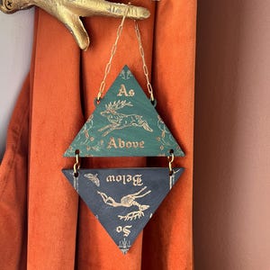 May include: A two-part hanging wall ornament featuring triangular wooden panels connected by gold chains. The top green panel displays a leaping deer and the text As Above, while the bottom dark panel shows a deer skeleton and the text So Below.