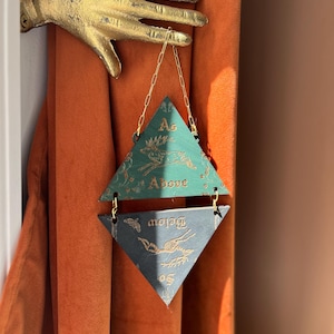 May include: A gold-colored metal hand wall mount holding a hanging ornament composed of two triangular wooden pieces connected by gold chains. The top green triangle features a gold deer illustration and the text As Above. The bottom dark triangle features a gold deer illustration and the text So Below.