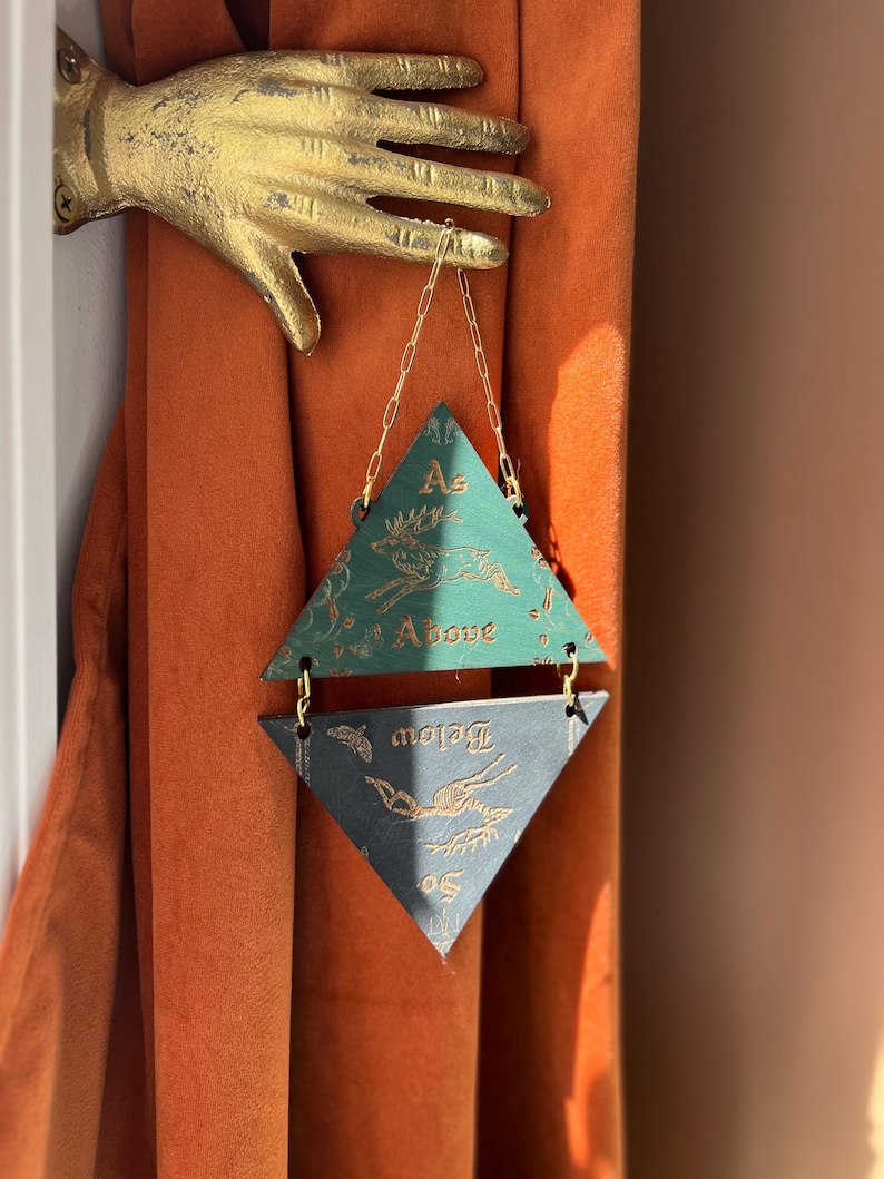 May include: A gold-colored metal hand wall mount holding a hanging ornament composed of two triangular wooden pieces connected by gold chains. The top green triangle features a gold deer illustration and the text As Above. The bottom dark triangle features a gold deer illustration and the text So Below.