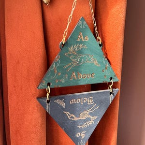 May include: A hanging wall ornament consisting of two triangular wooden panels connected by gold-colored metal chains. The top green panel features a gold deer illustration and the text As Above. The bottom dark panel features a gold deer skeleton and the text So Below.