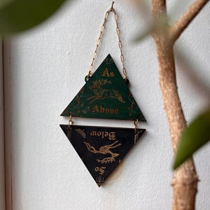 May include: A hanging wall decoration consisting of two triangular wooden plaques connected by gold metal jump rings and a chain. The top green triangle features a gold deer illustration and the text As Above. The bottom black triangle features a gold skeletal deer illustration and the text So Below.