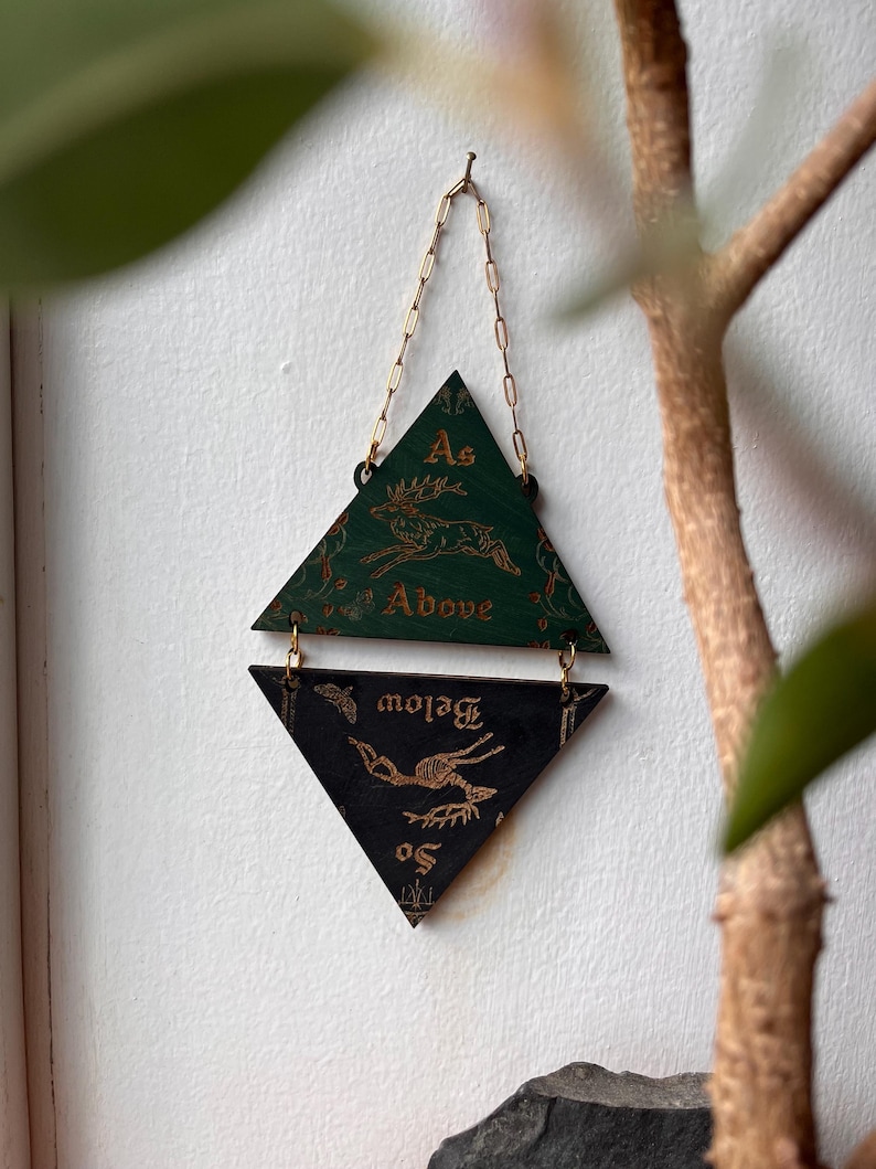 May include: A hanging wall decoration consisting of two triangular wooden plaques connected by gold metal jump rings and a chain. The top green triangle features a gold deer illustration and the text As Above. The bottom black triangle features a gold skeletal deer illustration and the text So Below.
