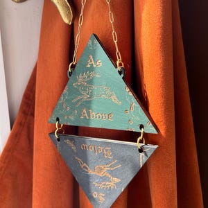 May include: A hanging wall decoration consisting of two stacked triangles connected by gold-colored metal chains. The top triangle is green with a gold deer illustration and the text "As Above". The bottom triangle is dark gray with a gold deer skeleton illustration and the text "So Below".