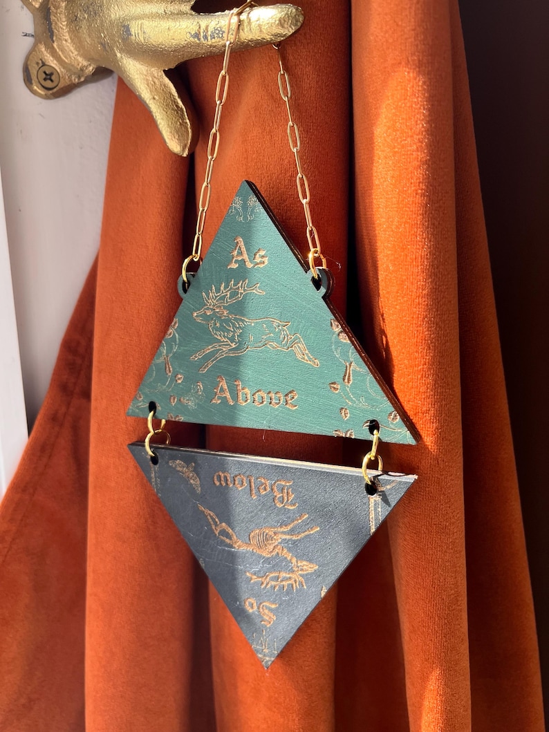 May include: A hanging wall decoration consisting of two stacked triangles connected by gold-colored metal chains. The top triangle is green with a gold deer illustration and the text "As Above". The bottom triangle is dark gray with a gold deer skeleton illustration and the text "So Below".