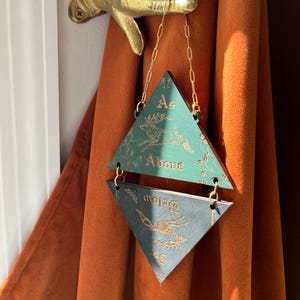 May include: A hanging wall ornament consisting of two triangular wooden plaques connected by gold-colored metal chains. The top green plaque features a gold deer illustration and the text "As Above," while the bottom black plaque features a gold deer skeleton illustration and the text "So Below."