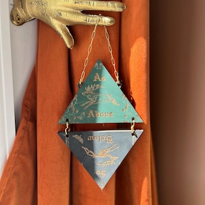 May include: A gold-colored metal hand wall mount holding a hanging ornament composed of two stacked triangles connected by gold chains. The top green triangle features a deer illustration and the text As Above, while the bottom dark triangle shows a skeletal deer and the text So Below.