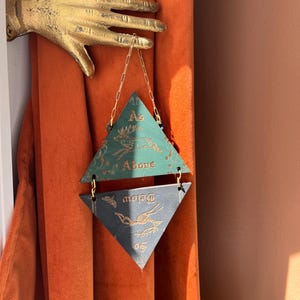 May include: A gold-colored wall-mounted hand sculpture holding a hanging ornament composed of two stacked triangles. The top green triangle features a deer illustration and the text As Above, while the bottom dark triangle features the text So Below.