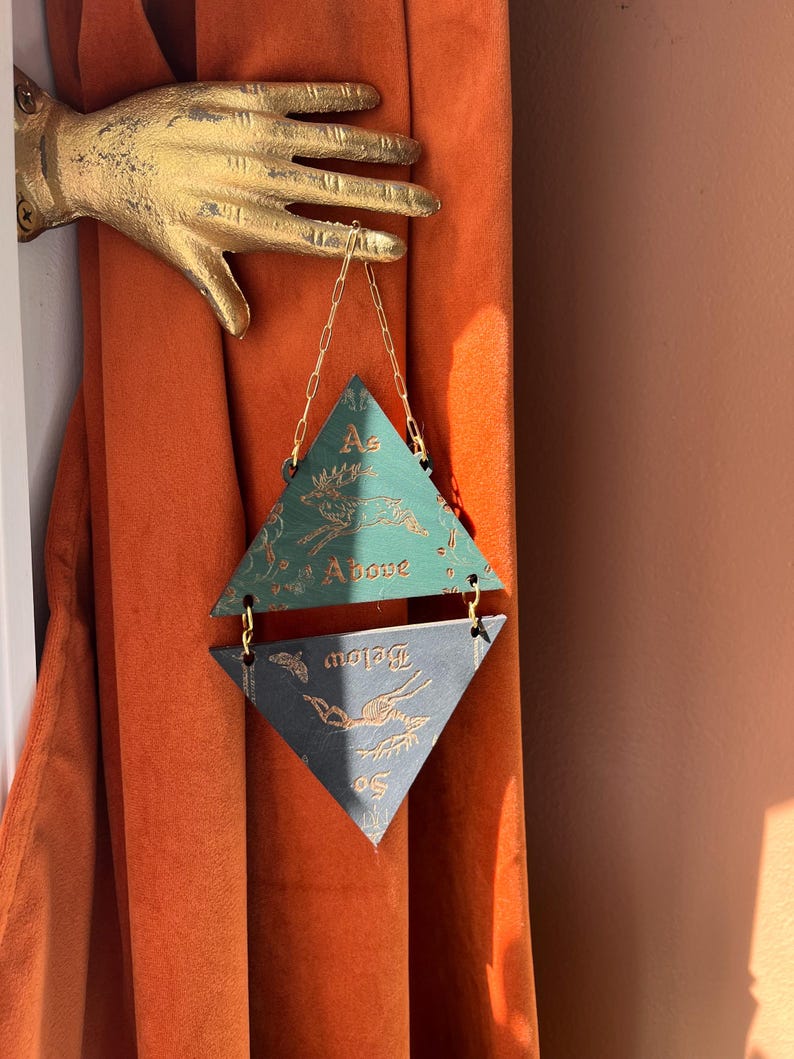 May include: A gold-colored wall-mounted hand sculpture holding a hanging ornament composed of two stacked triangles. The top green triangle features a deer illustration and the text As Above, while the bottom dark triangle features the text So Below.