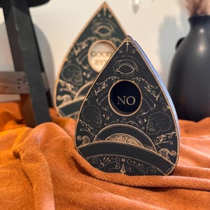 May include: A black wooden planchette featuring a central circular cutout with the word NO printed inside. The surface is decorated with intricate gold-toned line art depicting skulls, an eye, and ornate scrollwork patterns across the front.
