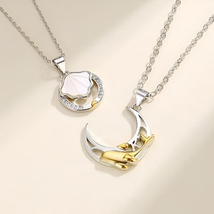 Puede incluir: Dos collares de plata con colgantes a juego. Un colgante es una luna creciente de plata con una mano dorada que sostiene un corazón. El otro colgante es un círculo de plata con una concha blanca en su interior.