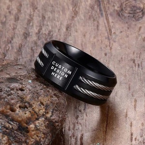 Op de afbeelding: Zwarte ring met een zilveren kabelontwerp en de tekst "CUSTOM DESIGN HERE" gegraveerd op de band.