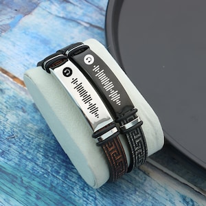 Puede incluir: Dos pulseras de cuero negro con detalles plateados. Cada pulsera presenta un icono de auriculares y un gráfico de onda sonora.