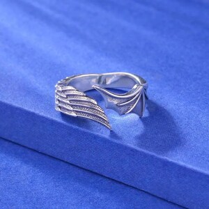 May include: A silver-colored ring with a wing design. One side features a feathered wing, while the other has a bat-like wing. The ring is set against a blue background.