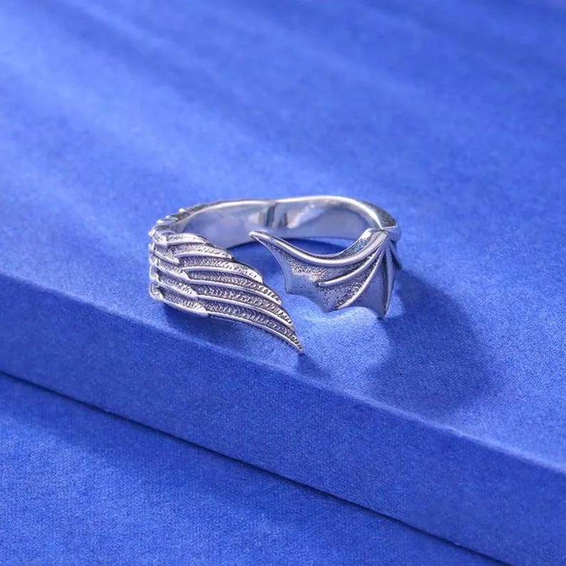 May include: A silver-colored ring with a wing design. One side features a feathered wing, while the other has a bat-like wing. The ring is set against a blue background.