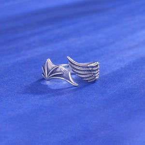 May include: A silver-toned ring with a wing design. One wing resembles a bat's wing, and the other wing has a feathered appearance. The ring is set against a blue background.