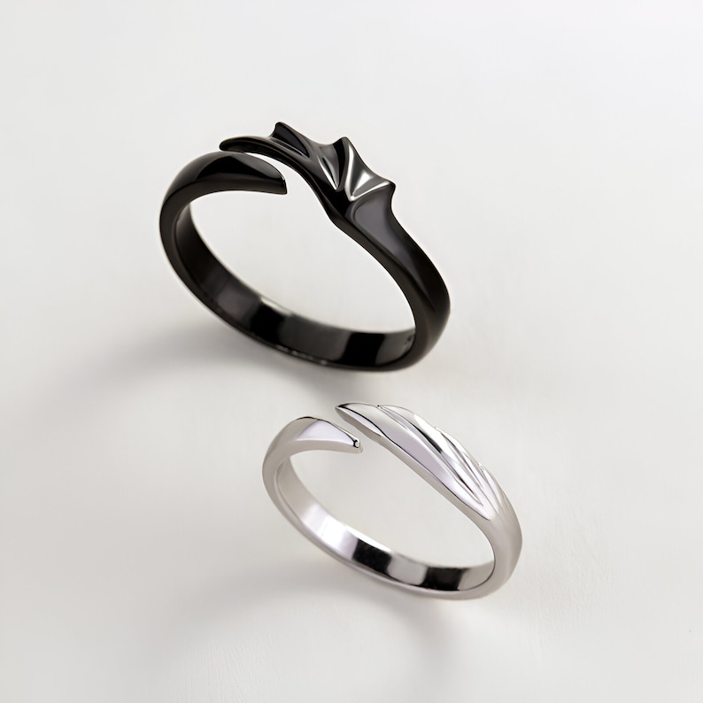 May include: A pair of matching rings, one black and one silver, with a unique design featuring a wing-like shape that interlocks when the rings are worn together.