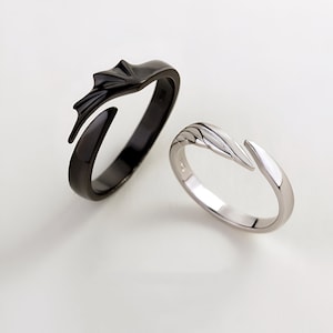 May include: A pair of matching rings, one black and one silver, with a unique design featuring a stylized wing shape. The rings are designed to fit together, creating a complete wing when worn as a couple's set.