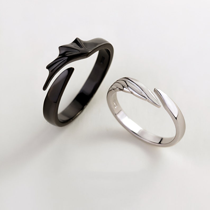 May include: A pair of matching rings, one black and one silver, with a unique design featuring a stylized wing shape. The rings are designed to fit together, creating a complete wing when worn as a couple's set.
