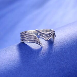 May include: A silver-colored ring with a wing design. One side of the ring features a detailed wing with layered feathers, while the other side has a bat-like wing shape. The ring is set against a blue background.