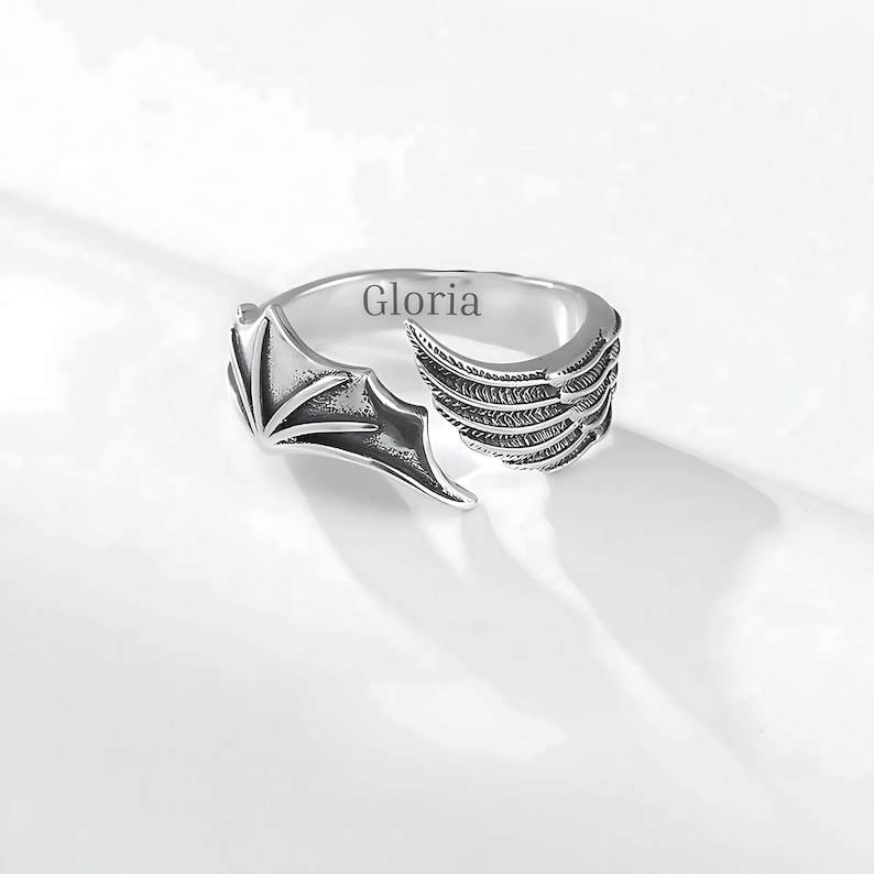May include: Silver-toned ring featuring a wing design. One side of the ring has a bat wing shape, while the other side has a feathered wing design. The word "Gloria" is engraved on the ring.