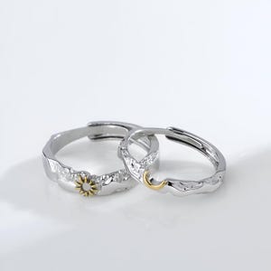 Puede incluir: Dos anillos de plata con bandas texturizadas. Uno presenta un pequeño diseño de sol dorado, y el otro una luna creciente dorada. Los anillos están sobre un fondo blanco.