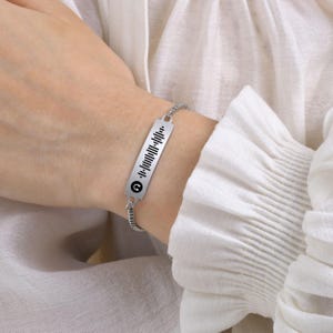Puede incluir: Una pulsera de plata con un dije rectangular que presenta un diseño de onda sonora en blanco y negro y un símbolo de nota musical negro.
