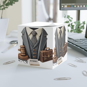 May include: A white cube-shaped notepad featuring a design of a gray suit jacket, tie, and a gavel. A brown briefcase and the word "ATTORNEY" are also displayed. The notepad is on a white desk with paperclips.