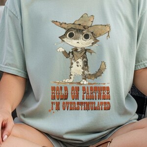 May include: A pale blue t-shirt with a cartoon cat in a cowboy hat and bandana. The cat has big eyes and is in a standing pose. The text "HOLD ON PARTNER I'M OVERSTIMULATED" is printed below the cat in a rustic font.